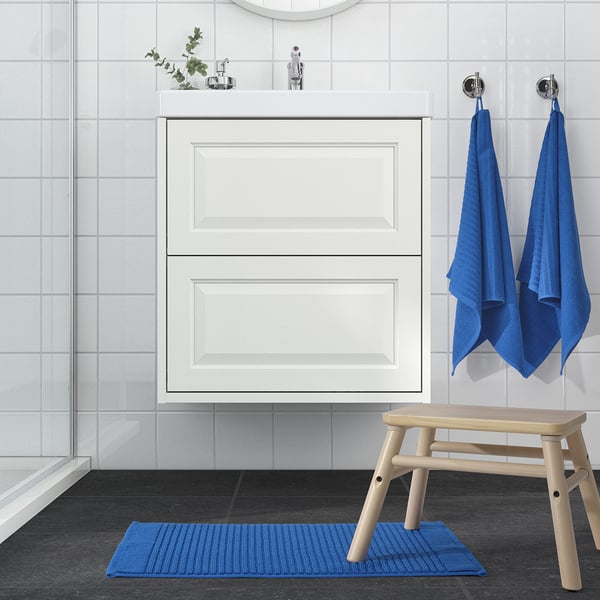 Modern bathroom with white cabinet, glass shower, blue ALSTERN mat and towels, light wood stool, grey floor tile, sleek decor.