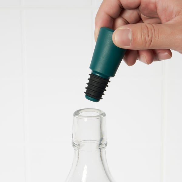 Hand placing green stopper into glass bottles neck. Prevents leakage, preserves taste.