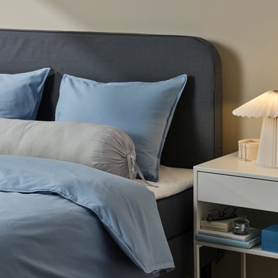 Grey BÄRGLIM bolster on bed with blue/gray bedding, dark grey headboard, white bedside table, lamp, glasses, and books. Recycled polyester pillowcase.
