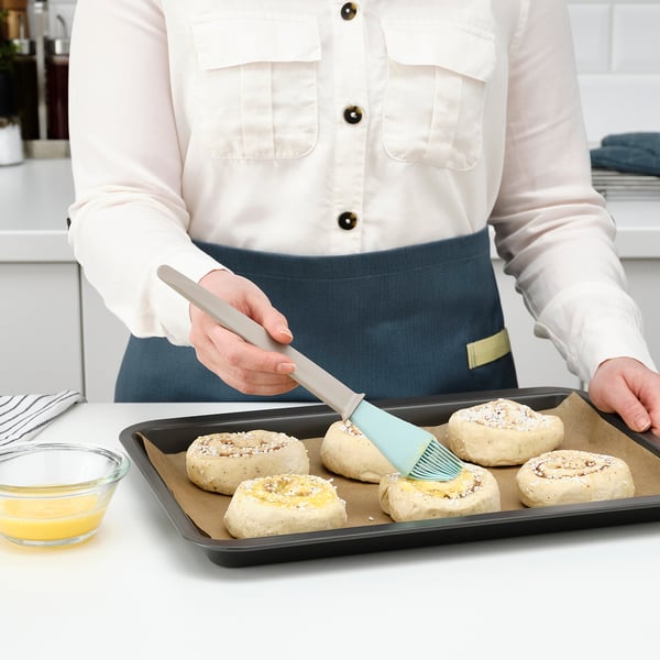 Person wearing an apron holds a brush, spreading liquid on pastries on a tray.