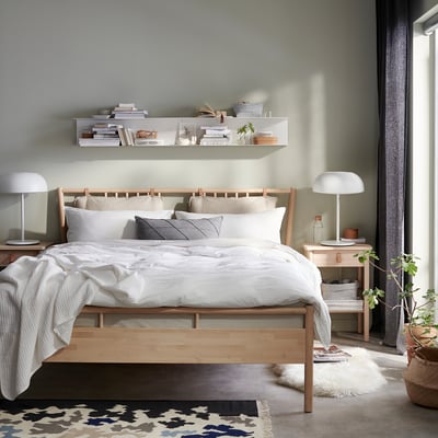 Cosy bedroom: light wood BJÖRKSNÄS bed with white linens, grey pillows, bedside lamps, and wall shelves with books and plants.