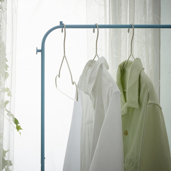 Clothes rack with three hangers: one green jacket, two white shirts, and hooks for accessories. Light, organised bedroom or hallway setup.