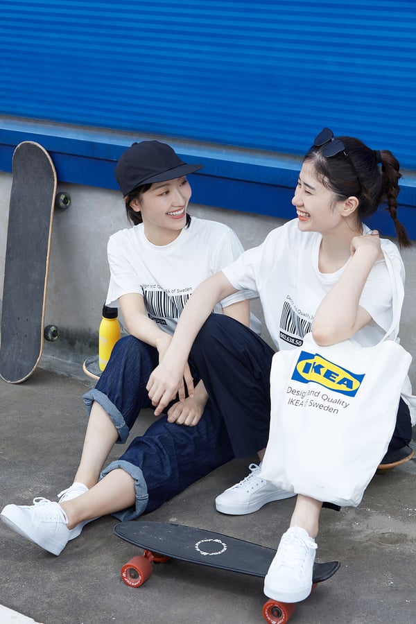 Two women sit on a skateboard, chatting. One holds an ikea white cotton tote.