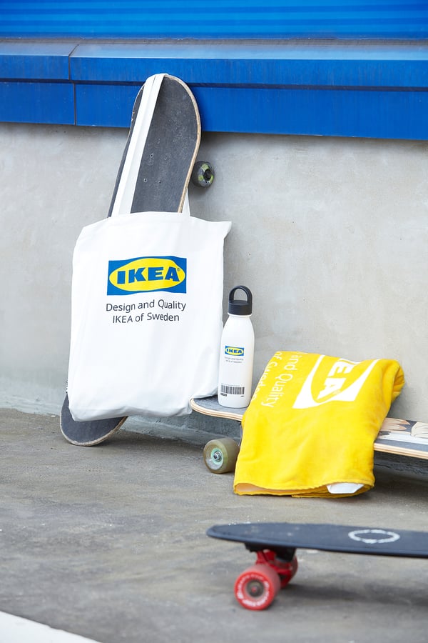 Ikea EFTERTRÄDA white tote bag with blue logo on a skateboard, alongside a yellow ikea bag and a water bottle by a blue and concrete wall.