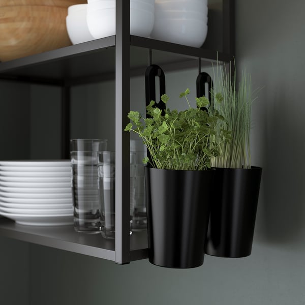 Black kitchen rail with hooks holding pots, one with fresh herbs, under a shelf with glasses and bowls.