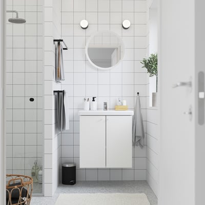 Modern white bathroom with ENHET vanity, round mirror, grey rubbish bin, black towel rail, and shower. Clean, minimalist design.
