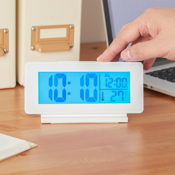 A hand is pressing a white digital alarm clock with blue backlight displaying time 10:01, alarm set to 12:00, and temperature 27°c.