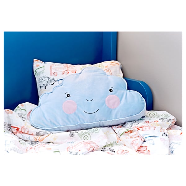 Blue cloud-shaped pillow with pink cheeks, smiling, on a playful bed with vehicle prints.