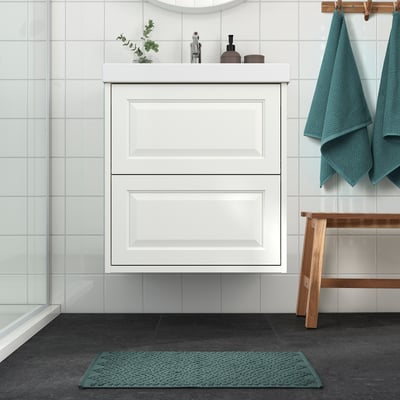 Modern bathroom with white cabinet, glass shower, wooden bench, and turquoise bathmat.