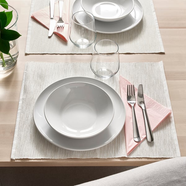 Two place settings with white plates, glasses, forks, knives, and pink napkins on woven placemats on a wooden table.
