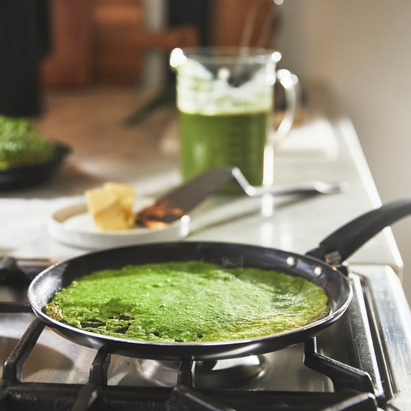 Green pancakes cooking in a non-stick frying pan on a stove, with a glass mug of green smoothie and food nearby.