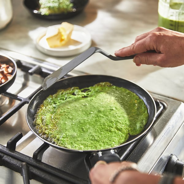 A person cooking green pancake batter in a non-stick pan on a stove, using a spatula.