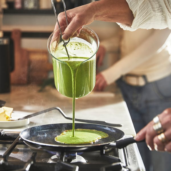 A person pours green batter from a blender into a non-stick frying pan on a stove, illustrating even heat distribution and easy food handling.