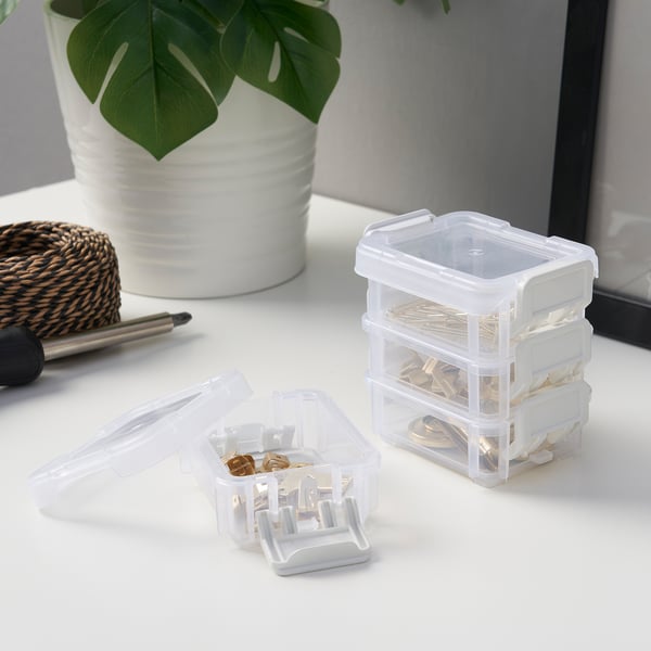 Clear plastic storage boxes stacked, one open showing small items inside, on a white desk with a plant and pen.