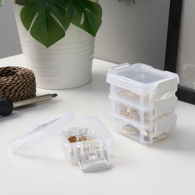 Clear plastic storage boxes stacked, one open showing small items inside, on a white desk with a plant and pen.