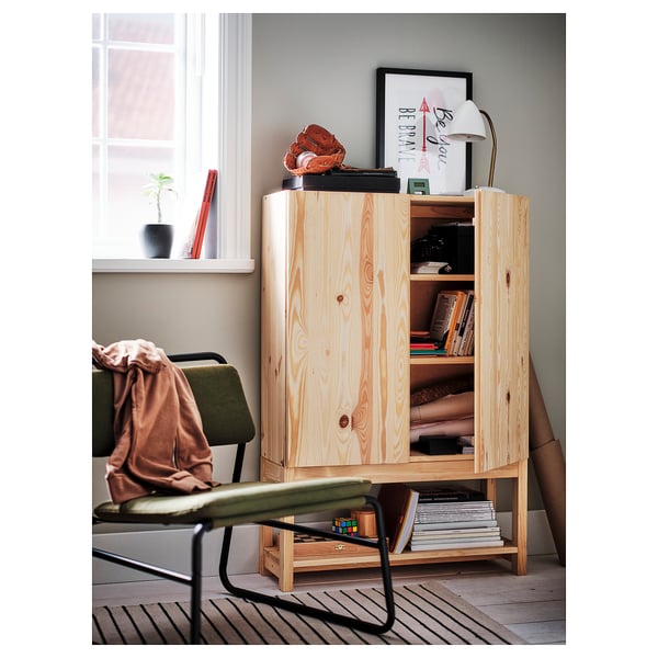 Light wood IVAR cabinet with open and closed storage, top display with lamp, books, and baseball mitt, next to green chair.