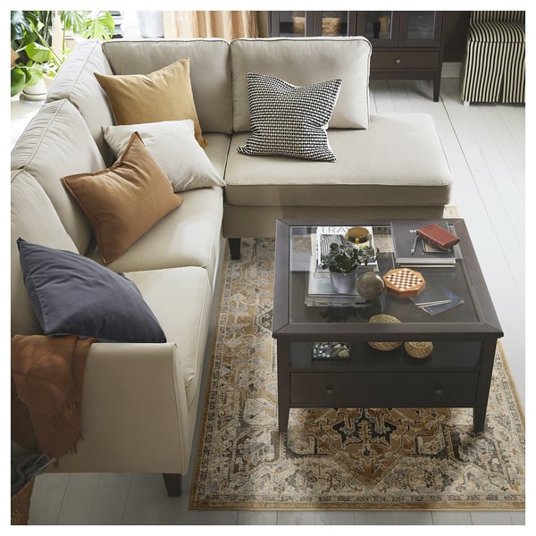 Large cream sectional sofa with colourful pillows, dark coffee table with various items atop, patterned rug beneath, set against light wooden floor.