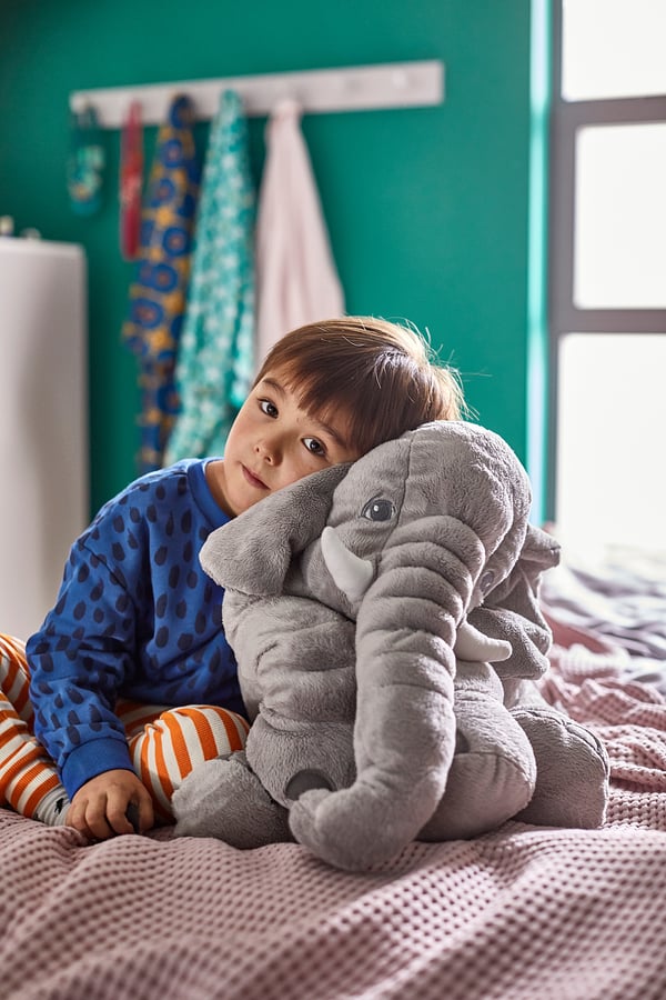 Child sitting on bed hugging large grey JÄTTESTOR elephant cuddly toy. Blue polka dot pyjamas, striped socks. Cosy, green-walled room.