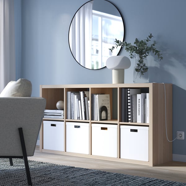 Ikea KALLAX light oak, square shelf unit holds books and decor, mirrored backdrop, grey upholstered chair with light blue wall.