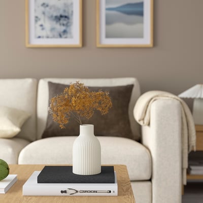 Modern living room with KAPKASTANJ vase on coffee table, yellow flowers, beige couch, and nature paintings.