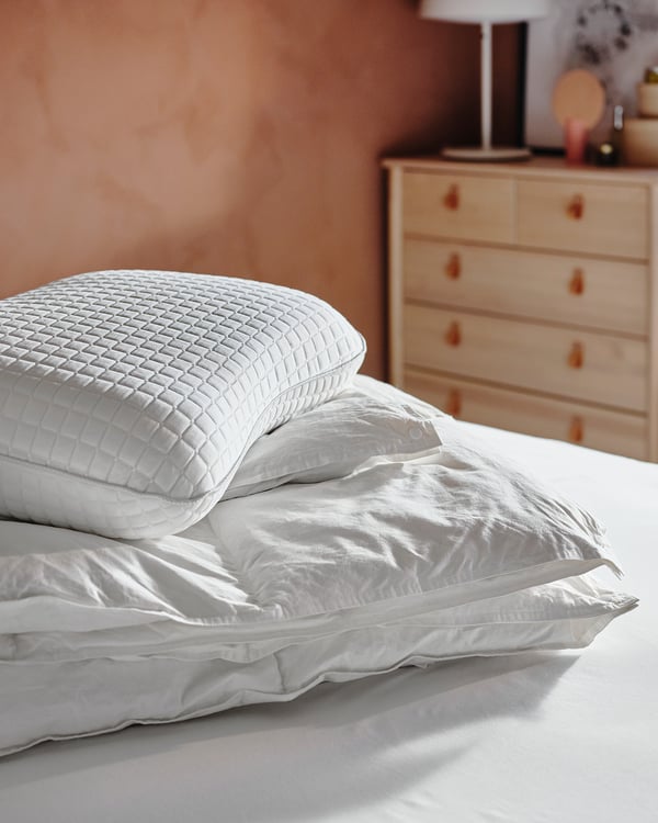Two white pillows on a bed, one with a quilted pattern and the other smooth. A light-colored wooden dresser with a lamp is visible in the background.