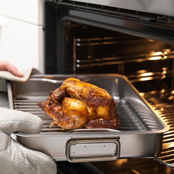 Person placing roast chicken in stainless steel roasting tin with a grill rack into an oven.
