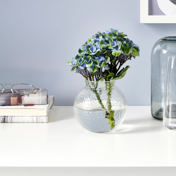 Clear glass vase with textured surface holds blue flowers on desk, surrounded by books and vases.