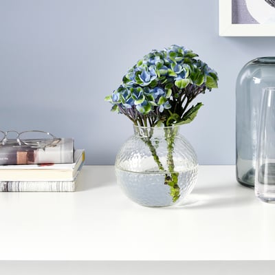 Clear glass vase with textured surface holds blue flowers on desk, surrounded by books and vases.