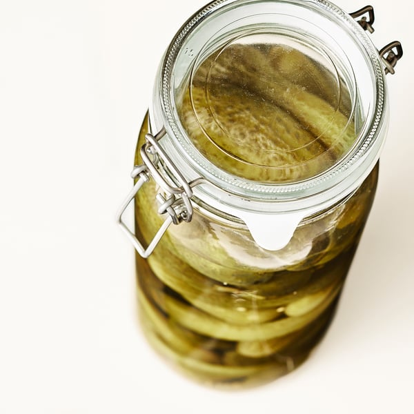 A clear glass jar with metal clasp, filled with pickles submerged in liquid. The airtight seal preserves freshness.