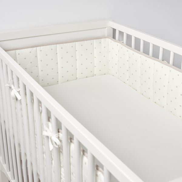 A crib with a white bumper pad that has small dots, tied with ribbons, ensuring baby safety and comfort by preventing bumps.