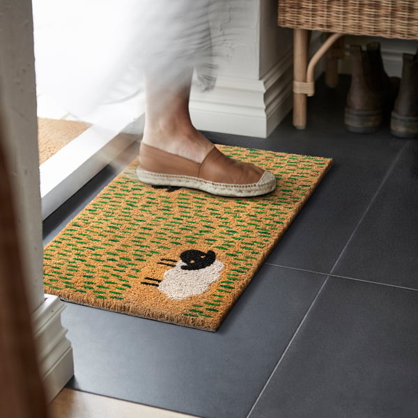 Person stepping on yellow-green door mat with sheep image, preventing dirt inside.
