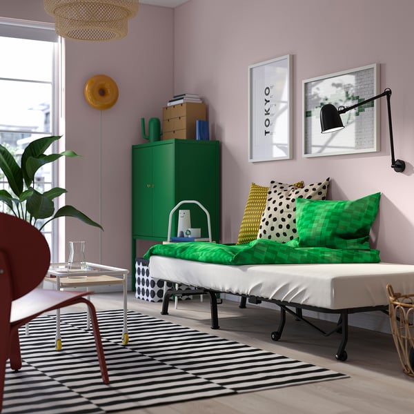 Bright guest room with green bed, black-and-white striped rug, red chair, green storage unit with books, and wall art.