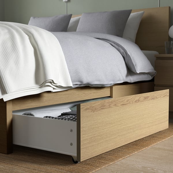 A wooden bed frame with open drawer under it revealing folded clothes; light grey bedding on top.