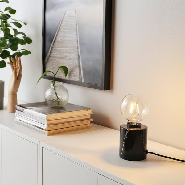 A sleek, black marfrost lamp with a marble base and a warmly lit led bulb on a white surface. Its surrounded by books and a small plant.