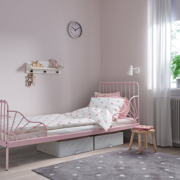 A pink MINNEN extendable metal bed in a kids room. It heart-patterned bedding and is larger than the nearby wooden stool.