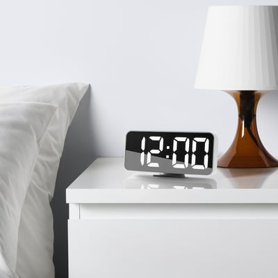White NOLLNING alarm clock with large digits sits on a bedside table. Beside it is a matching lamp and a bed with white sheets.