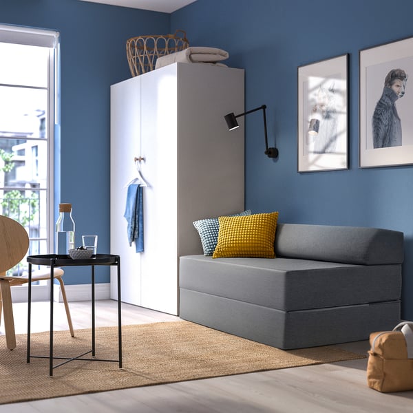 Modern bedroom with grey sofa-bed against blue wall, decorated with yellow and blue pillows, near a closet and a small table with wooden chair.