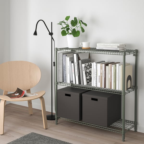 OMAR olive green storage unit with shelves holding books, mug, plant, and bins. Nearby, a wooden chair with a magazine and a black adjustable lamp.