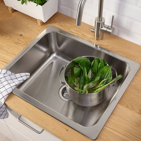Stainless sink with strainer full of green veggies, wooden countertop, modern tap.