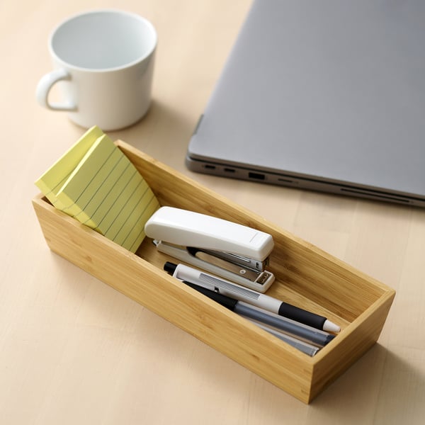 Wooden desk tray holding yellow sticky notes, stapler, and pens beside a laptop and a cup for organised workspace items.