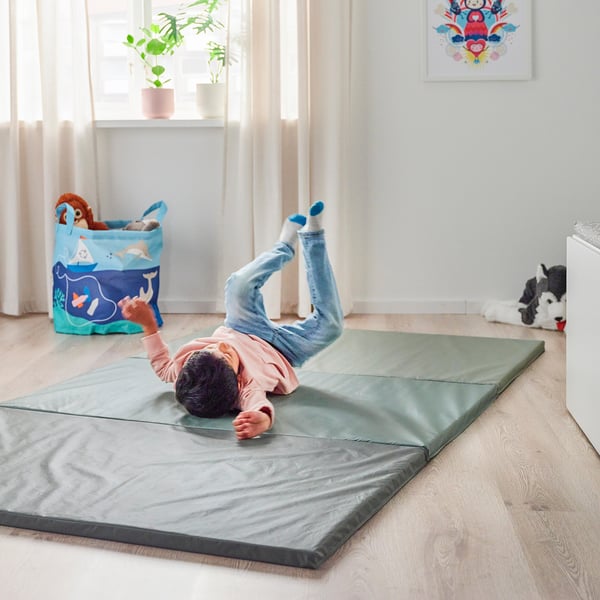 Baby playing on a soft, foldable PASSBIT gym mat, practising somersaults indoors. Nearby, a toy bin and stuffed animals are visible.