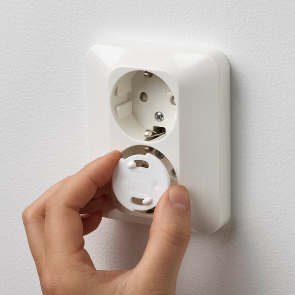 A hand inserts a white safety plug into a wall socket to prevent access, ensuring child safety.