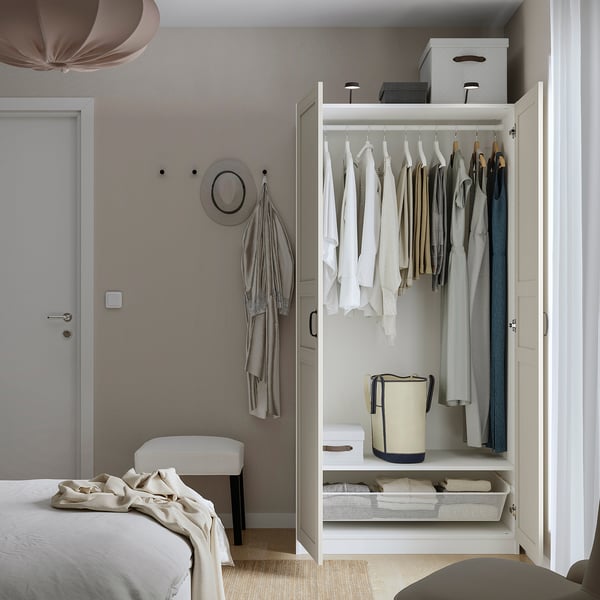Open wardrobe displaying clothes, storage bins, and a laundry basket. Shown in a minimalist bedroom with a bed and a stool.