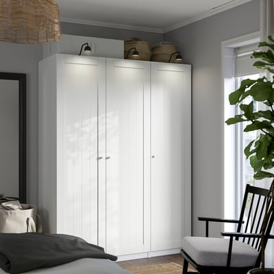 Large white PAX wardrobe, tall & wide, with lit top shelves. Surrounded by grey bed & black chair, green plant nearby.