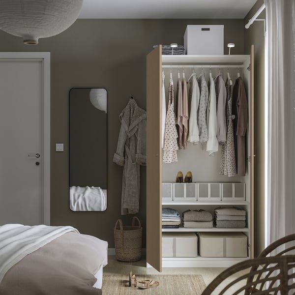 A modern bedroom with a PAX wardrobe open, displaying clothes, shelves with folded clothes, and storage bins. A mirror and a robe hang on the wall.