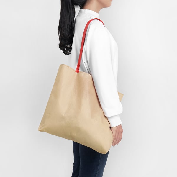 A person carries a large beige shopping bag over their shoulder with a red strap. The bag folds into a compact size for easy carrying.