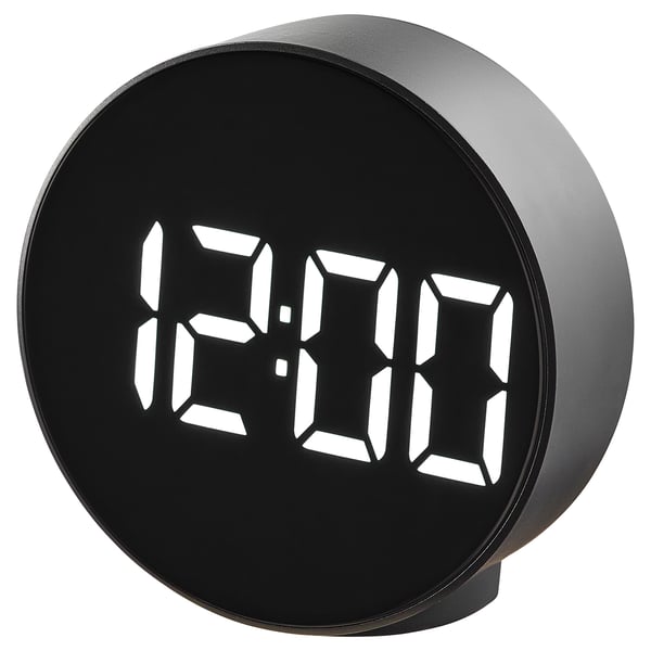A black round digital alarm clock, named PLUGGET, displays time in bright white digits. It stands on a silicone rubber pad.