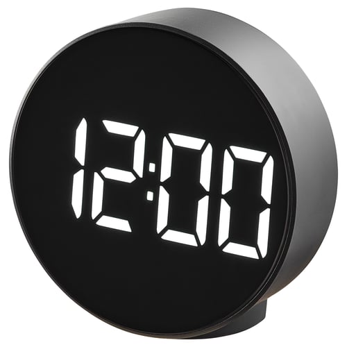 A black round digital alarm clock, named PLUGGET, displays time in bright white digits. It stands on a silicone rubber pad.