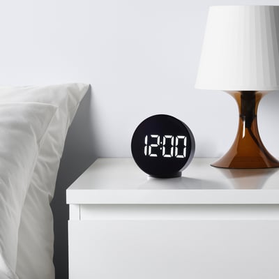 Modern black alarm clock on bedside table displaying 12:00, near minimalist bed with lamp.