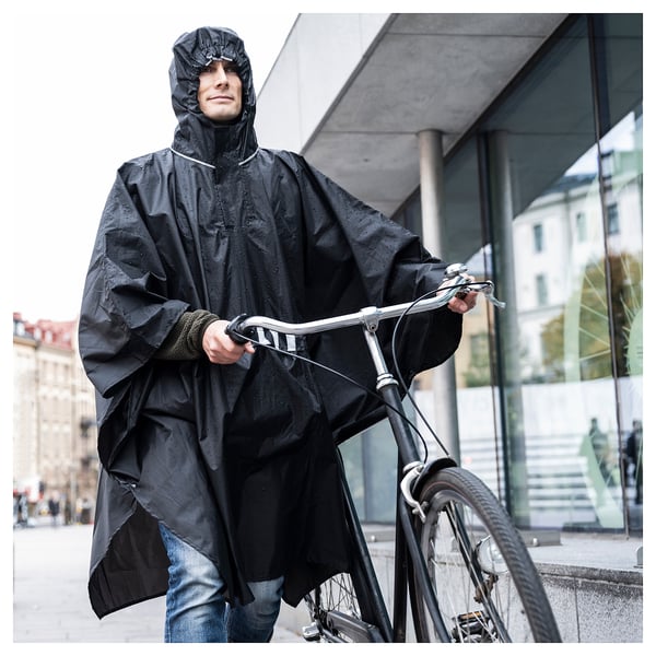 Person wearing black SALTSTEN rain poncho, holding bicycle. Urban setting, modern building in the background.